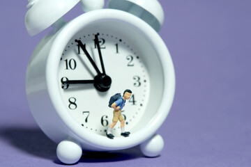 Miniature tiny people toy photography. A boy student with backpack standing on clock. Isolated on purple background