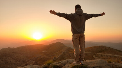 Fototapeta na wymiar Man standing with open arms on a mountaintop. Person standing on the cliff edge facing the rising sun. Scenery sunrise in mountainside. Embracing the world