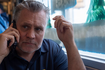 portrait of a middle aged man gray hair and facial hair making a phone call with stern expression and hand gesturing sitting down to eat at a diner wearing a dark blue polo sat in front of the window