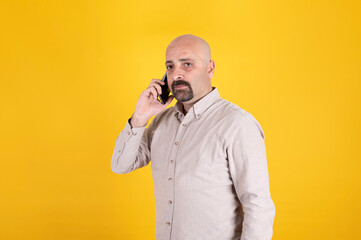 Middle aged man talking on the phone, standing over yellow studio background. Bald male seriously...