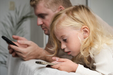 Girl and adult man lying on bed using phones at home. Child and father looking at gadgets, spending time together.