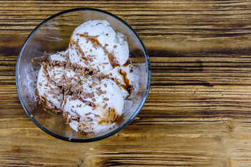 Glass bowl with ice cream balls and chocolate topping on a wooden table. Top view