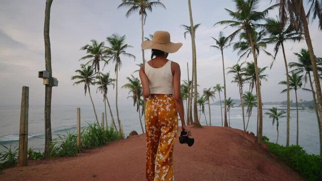African american woman walking with camera on tropical island. Female multiracial photographer tourist explores ocean view location on vacation. Black girl in straw hat taking pictures on sunrise.