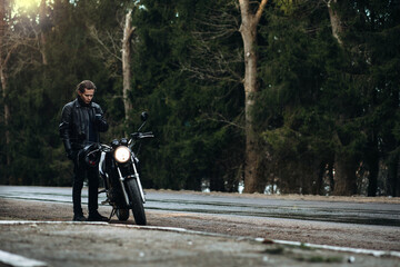 male motorcyclist in a leather motorcycle jacket stands by a motorcycle on a forest road....