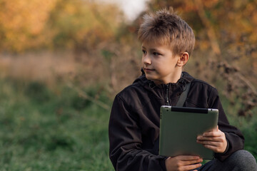 A portrait of a European blond boy in a black jacket with a tablet outside a school looking to the side for online learning, a happy young man in a park. A happy schoolboy. Autumn photo, front view