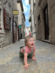 Beautiful Tourist Baby In Love Walking Alone in Old city Street. Happy Young Kid Toddler Smiling Walking Around European Town Streets, Looking At Architecture. Travel Concept. High Quality Image.