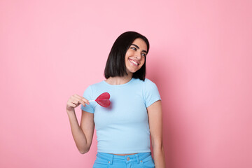 Happy young woman holding heart shaped lollipop over eye against pink background