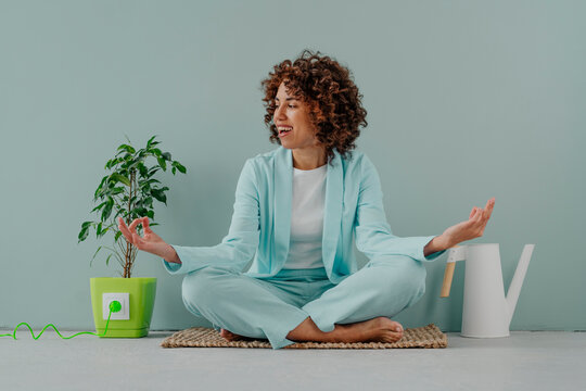 Happy woman meditating by potted plant and watering can in front of blue wall