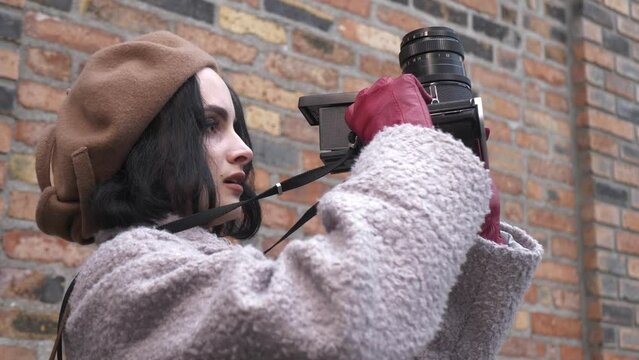 Video portrait. A young woman in a beret on the background of a brick wall on a city street takes pictures with an old film camera. Stylish young lady. Creative person