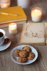 Cup of tea or coffee, plate of cookies, books, e-reader, pencil and lit candles on the table. Selective focus.