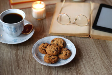 Cup of tea or coffee, plate of cookies, books, e-reader, pencil and lit candles on the table. Selective focus.