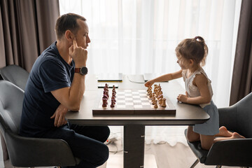 Little girl playing chess with her father at the table in home kitchen. The concept early childhood development and education. Family leisure, communication and recreation.