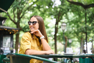 Charming brunette woman in sunglasses and yellow shirt spending time on the terrace of cafe. Carefree serene young woman enjoying morning coffee outdoors