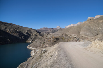 Dusty road in the autumn mountains on a sunny November morning on a slope, at the bottom of the slope there is a blue mountain lake