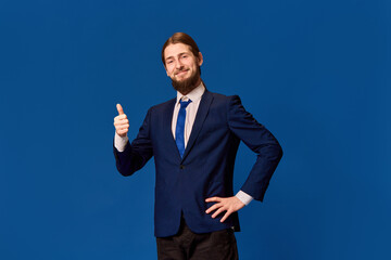 Approval. Portrait of happy young man in formal wear rising finger with pleasant gesture against blue studio background. Good job. Concept of emotions, facial expression, lifestyle, occupation