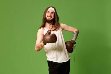 Portrait of young man with long brown hair posing in singlet and leather boxing gloves against green studio background. Concept of emotions, facial expression, lifestyle