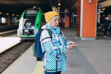 Old happy woman with backpack in yellow hat waiting train on station platform and using smart phone on urban background. Railroad transport concept