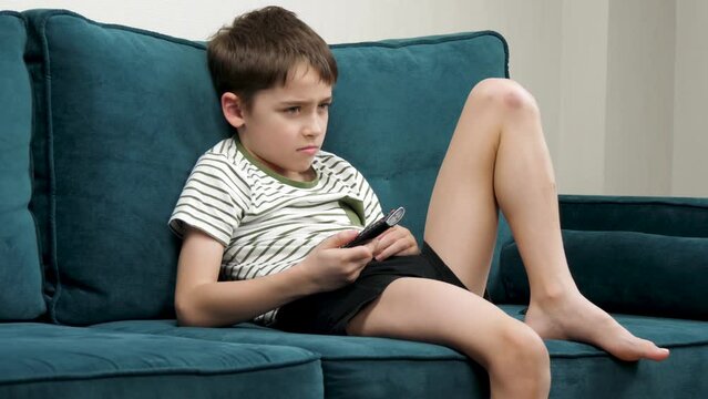 A charming child is sitting on the sofa in the room and watching TV, pressing the buttons of the remote control. Childrens leisure