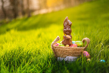 Easter eggs in basket with easter bunny on top. Chocolate rabbit with colorful decorated eggs in wicker basket in grass. Magical morning light, spring season holidays. Traditional egg hunt