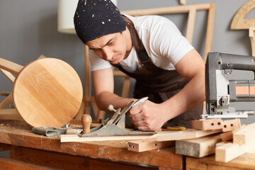 Indoor shot of concentrated young carpenter wearing apron and cap working in workshop, using plane,...