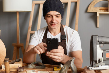 Indoor shot of handsome caucasian young adult man in cap and apron using his mobile phone in workshop, having break, chatting with friends, checking social networks.