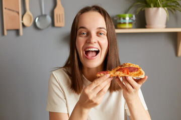 Indoor shot of happy cheerful woman eating delicious piece of pizza, holding fast food snack, looking at camera with amazed face, wearing casual white T-shirt posing in kitchen