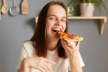 Portrait of carefree laughing woman with brown hair biting piece of pizza, feels awesome wearing white T-shirt posing in kitchen, enjoying junk food for dinner.