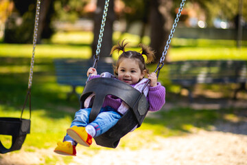 Cute caucasian little toddler girl with two tails smiling widely and swinging on a baby swing on the playground with violet sweeter sleeveless jacket and jeans