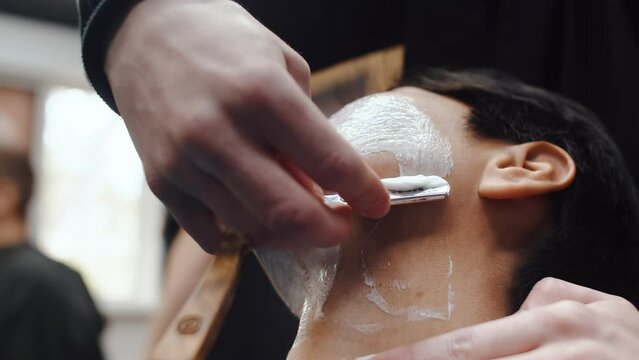 Professional barber slowly and carefully shaves a male client's neck with a straight razor using a shaving foam close up. Man being shaved in a barbershop