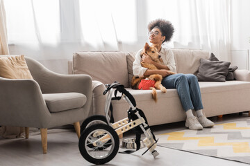Curly african american woman in eyeglasses hugging dog with special needs near wheelchair in living room.