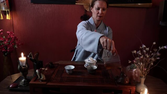 female master in kimano conducts a tea ceremony and pours hot tea on figurines while sitting at a table, authentic tea paraphernalia