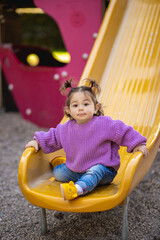 Fototapeta na wymiar Sweet caucasian little toddler girl with two tails having fun sitting on the yellow hill in the playground with violet sweeter sleeveless jacket jeans