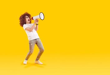 Cheerful funny preteen boy with loudspeaker in his hands makes loud advertisement on orange background. Full length child boy in curly wig announces children's sale with megaphone. Copy space. Banner. - obrazy, fototapety, plakaty