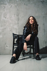 Plakat A cute young woman in a leather jacket and black pants wearing sunglasses is sitting on a chair on a gray background.