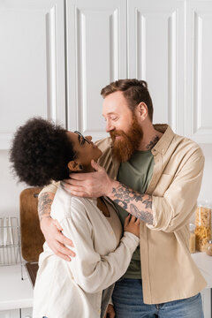Positive tattooed man touching african american girlfriend in kitchen at home.