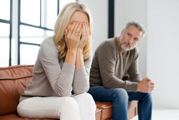 Fototapeta na wymiar Upset middle-aged couple is arguing at home, frustrated blonde woman covered face with palms and crying, sadness grey-haired mature man on background. Spouses have difficulties, relationship crisis