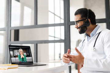 Indian male doctor with a stethoscope in gown sitting at the desk and looking at the laptop, giving consultation to patient online and discussing the insurance plan. Telemedicine concept