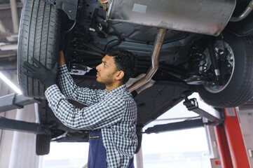 Indian car mechanic standing and working in service station. Car specialists examining the lifted car. Professional repairmen wearing mechanic uniform in blue color.