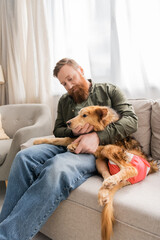 Bearded man hugging handicapped dog while sitting on couch at home.