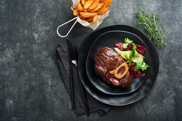Steaks Osso Buco. Sliced beef grilled Osso Buco or porterhouse meat steak with garnished with salad and french fries on black marble board on old wooden background. Top view. Mock up.