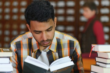 Portrait of cheerful male international Indian student with backpack, learning accessories standing near bookshelves at university library or book store during break between lessons. Education concept
