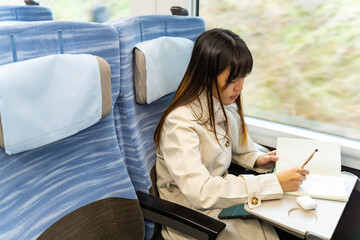  Asian woman writing journey memories in diary book during travel on railway transportation in winter vacation. Businesswoman working outside office. Work from anywhere and solo travel concept.