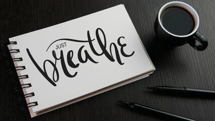 JUST BREATHE black lettering in notebook with cup of coffee and pens on black wooden desk