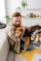 Bearded man with tattoo hugging disabled dog in wheelchair at home.