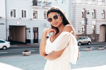 Fototapeta premium Young beautiful smiling hipster woman in white dress. Sexy carefree model posing on the street background at sunset. Positive female outdoors at warm sunny day. Cheerful and happy. In sunglasses