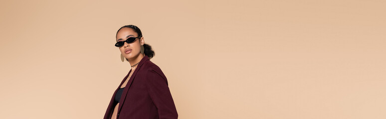 stylish african american woman in burgundy blazer and sunglasses looking at camera isolated on...