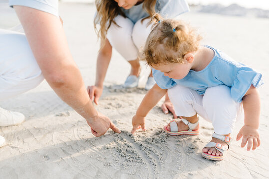 Happy young family with a little daughter girl on the sand together outdoors. Mother, father, and child playing on the sandy beach. Family day and childhood concept.