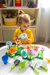 Fototapeta premium A little girl playing with colorful flower garden toys and planting tools on white table. Garden themed toys for kids. Pretend play, Montessori activity
