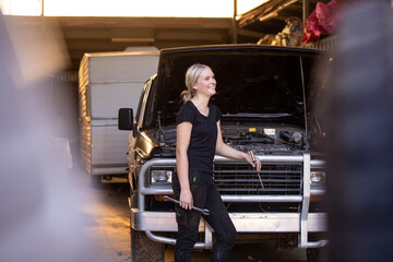 Young female aussie mechanic holding tools and leaning on a car in need of repair in workshop garage