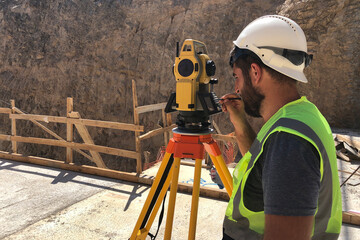 Land surveyor stands by theodolite on a tripod against construction site at background. Topographer...